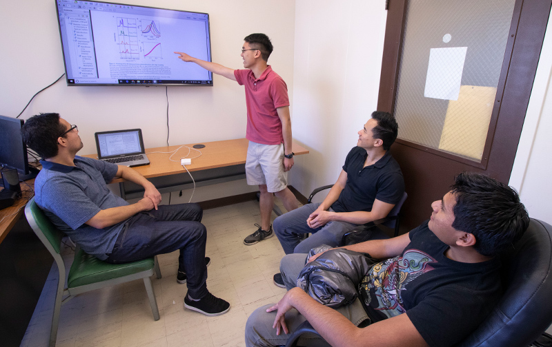 Graduate student Matthew Du points to the “pink line,” indicating exciting results from the study. His colleagues Raphael Ribeiro (left), Joel Yuen-Zhou (top right) and Luis Martínez-Martínez look on. Photo by Michelle Fredricks, UC San Diego Physical Sciences