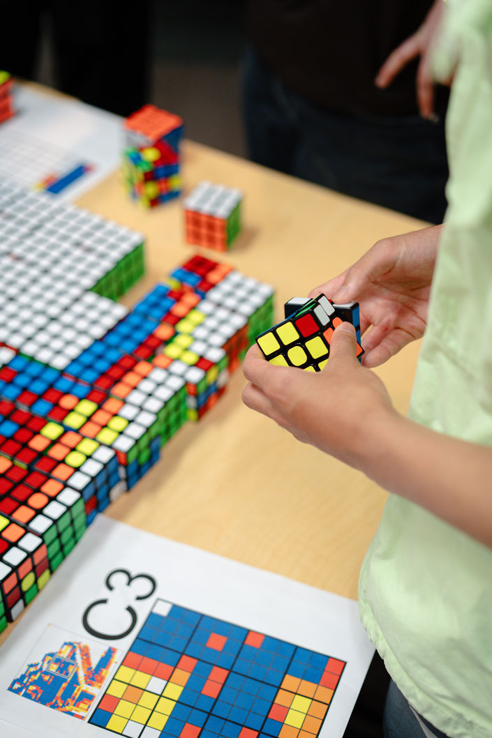 Hands hold a Rubik's cube mid-twist, with a number of Rubik's cubes on the table in front shown in various combinations and a piece of paper designated as C3 showing patterns.