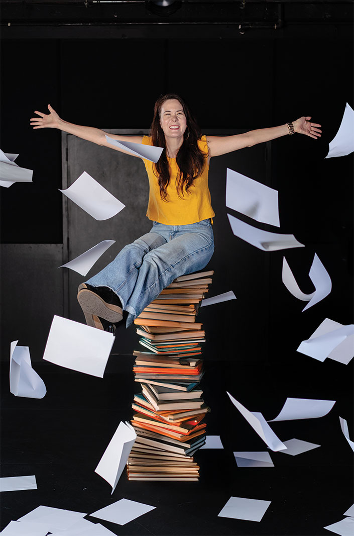 A woman sits atop a stack of books with her arms stretched wide, pieces of paper falling in the air around her.
