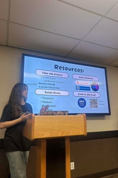 A person stands at a podium with a powerpoint slide showing on a screen behind them that has the header: Resources.