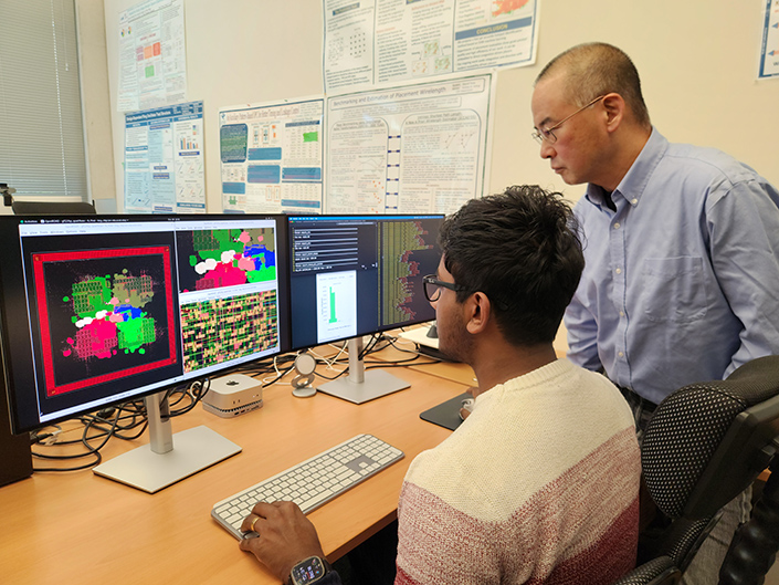 Two people in front of two computer screens: one displaying multi-colored patterns, the other displaying code.