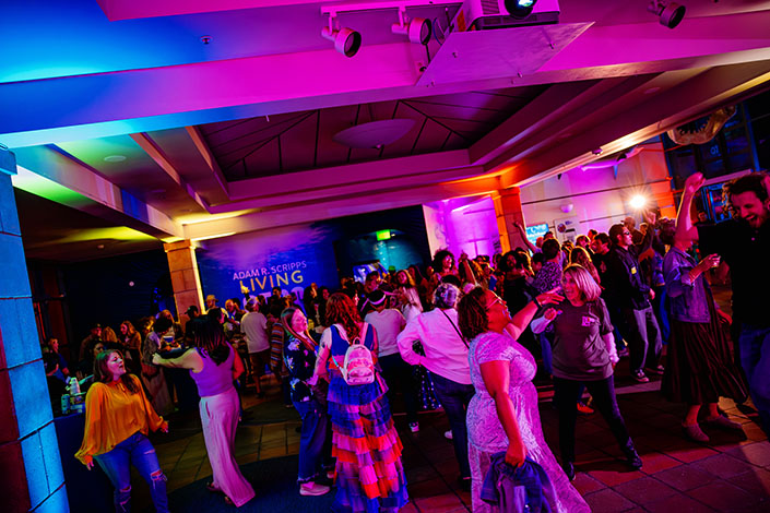 A crowd of people dance in a large room illuminated by pink and blue lights.