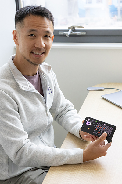 Man in a light gray sweater sits at a desk while holding a smartphone.