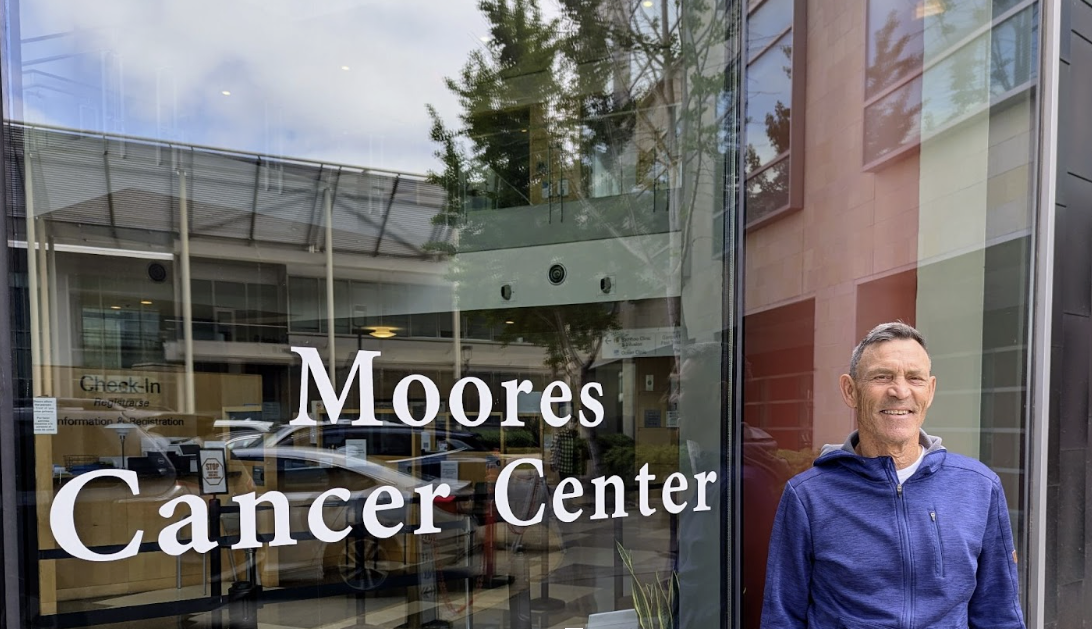 Photo of a man in a blue windbreaker in front of a window that says Moores Cancer Center