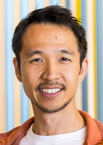 Portrait of a man in a white shirt and orange jacket against a background patterned with yellow and blue vertical stripes.
