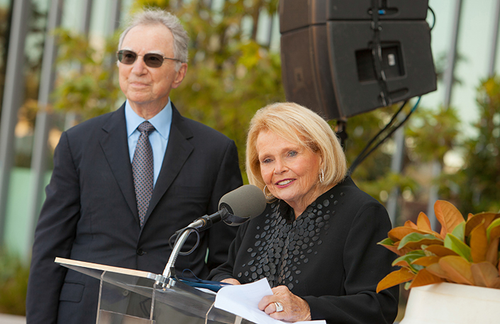 Joan Jacobs speaking at a podium, with Irwin Jacobs in the background