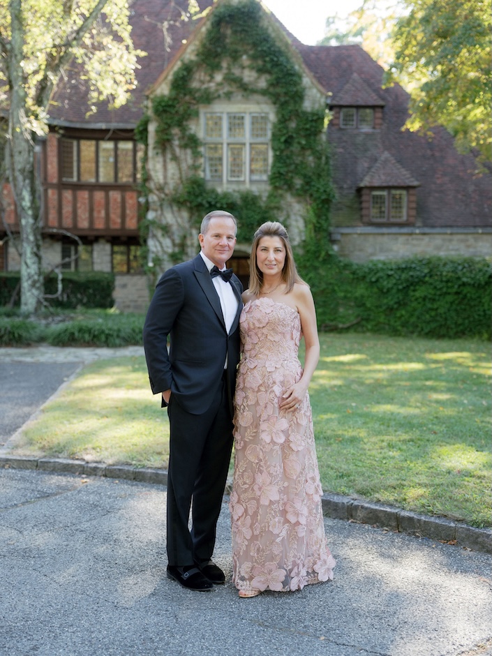 Man in tux and woman in beige ballgown in front of Victorian style home with vines growing up it.
