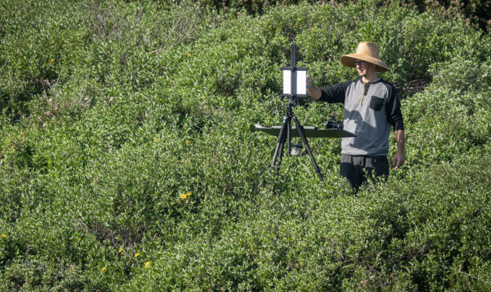 A man in a straw sunhat stands among the brush and greenery with an easel, canvas and paints.