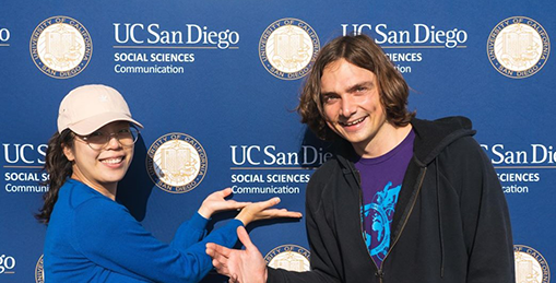 Two Communication students gesture at wall decorated with department/school logos