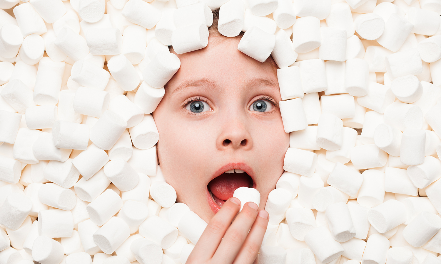 Child surrounded by marshmallows about to eat a marshmallow