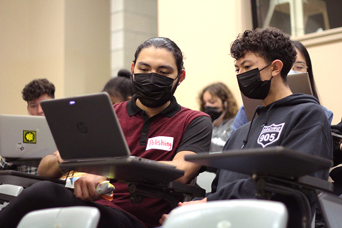 A UC San Diego undergraduate student sits in a lecture hall on campus next to a high school student, looking at a laptop and helping him craft his personal insight question answer.