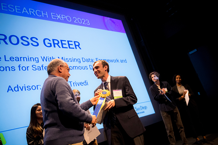 A man in a blue sweater hands a man in a suit a gold and blue first place ribbon.