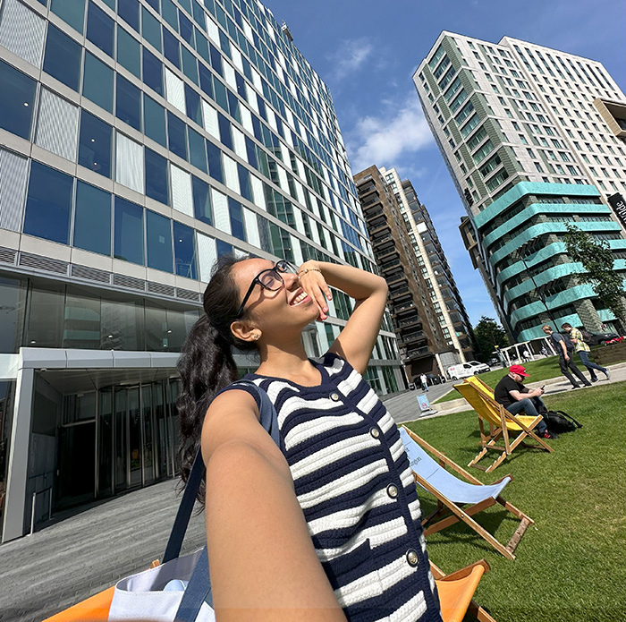 A student takes a photo of herself against a backdrop of buildings on a sunny day.