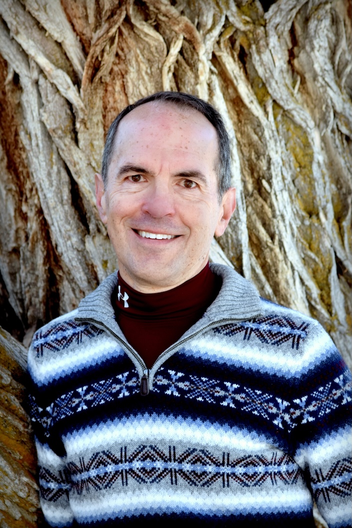 Picture of man with a sweater against a tree trunk, smiling