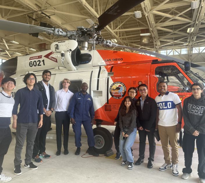 Military officer and nine students stand in front of a specialized helicopter.