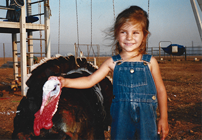 A young girl in overalls pets a turkey in her backyard.