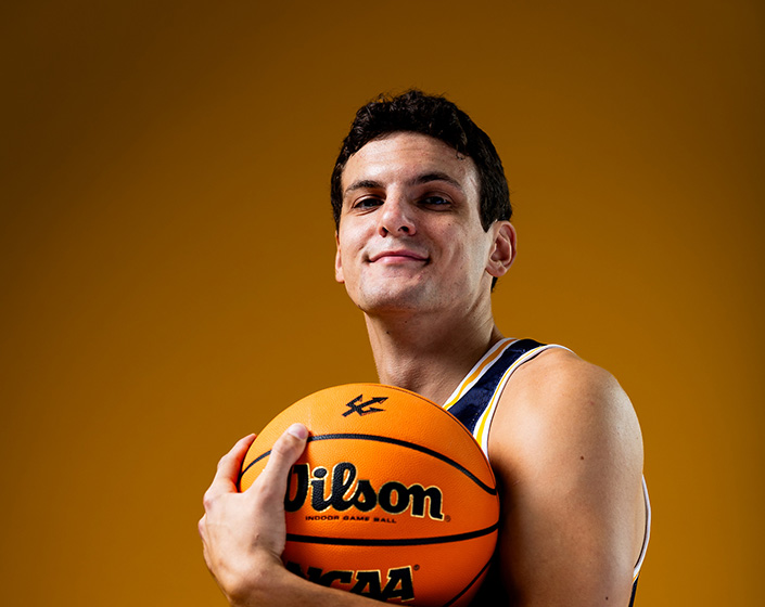Tyson Dunn cradles a basketball in his left arm and looks down at the camera. Tyson wears the UC San Diego Basketball jersey and appears against a brown background.