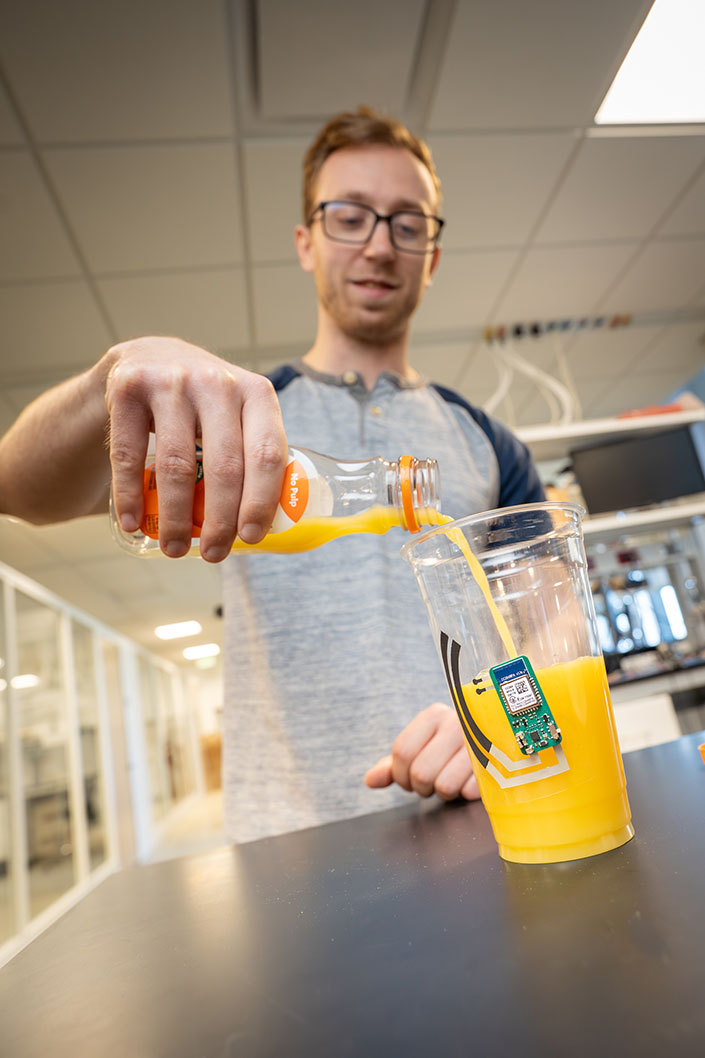 Person pours orange juice into a clear cup, which has a small circuit attached on the outside.