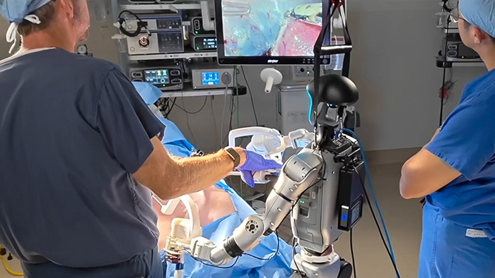 A surgeon stands near a humanoid robot. Another surgeon stands nearby and looks at a screen showing the surgery. 