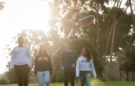 A group of four UC San Diego students walking on campus next to the Sun God statue with the sun shining behind them.