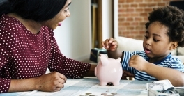 Black mom and young son at kitchen table with son dropping coins in piggy bank