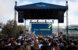 Hundreds of students sit in rows of chairs facing a large stage with a speaker at the podium and video screen behind that says 