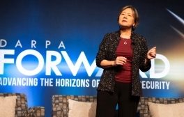 DARPA Director Stefanie Tompkins gestures with one hand while standing on a stage in front of a projection of the DARPA Forward, Advancing the Horizons of National Security logo.