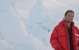 David Lesser stands against an icy Antarctic backdrop wearing a heavy red jacket.