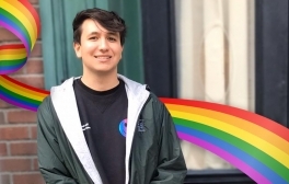 Isaiah LeDonne smiles while standing next to a building. He has a rainbow graphic behind him.