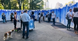 People mill about in front of a large white canvas with blue lines painted on it