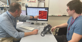 Researcher Chris Holland sits behind desk and points at computer monitor as student watches.