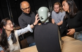 Students and a male professor gather around a test dummy used for audio research.