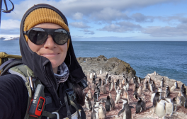 A selfie of a woman researcher who is in the field in Antarctica. She is surrounded by chinstrap penguins.