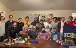 The UC San Diego Men's Club Water Polo team poses for a group photo in a living room after winning a national title, smiling and holding a UC San Diego flag.