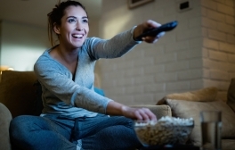 Young happy woman changing channels with remote control while watching TV and eating popcorn in the evening at home.