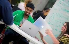 Two students sit at a table at the Teaching + Learning Commons, each with a computer, smiling as they collaborate on a project. A board with math equations is in the background.