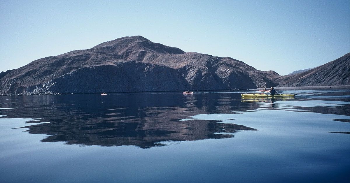 A kayaker floats on water that is so calm it reflects the surrounding mountains.