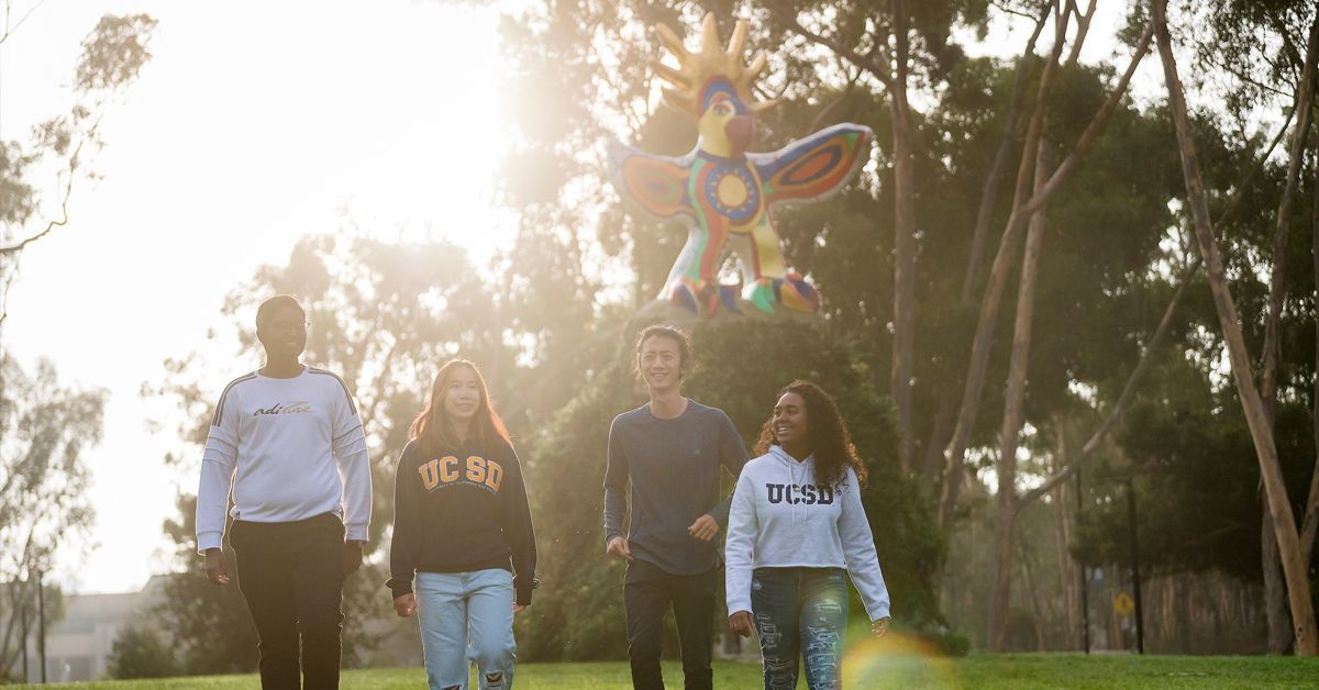 A group of four UC San Diego students walking on campus next to the Sun God statue with the sun shining behind them.