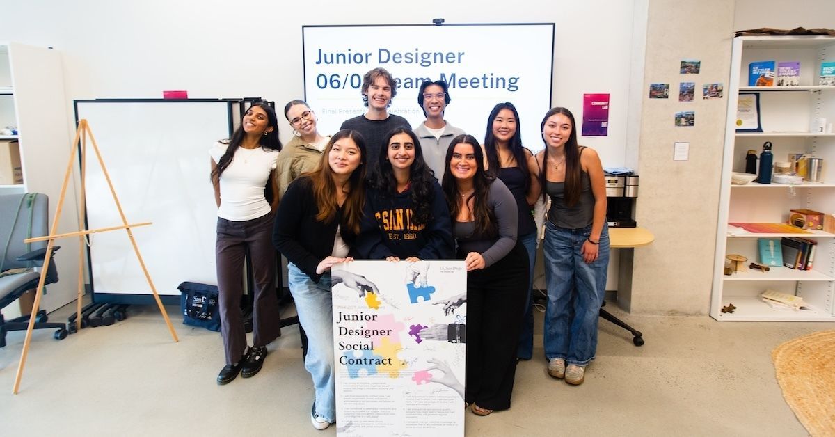 Nine students pose in a classroom environment. They are holding a poster that says 