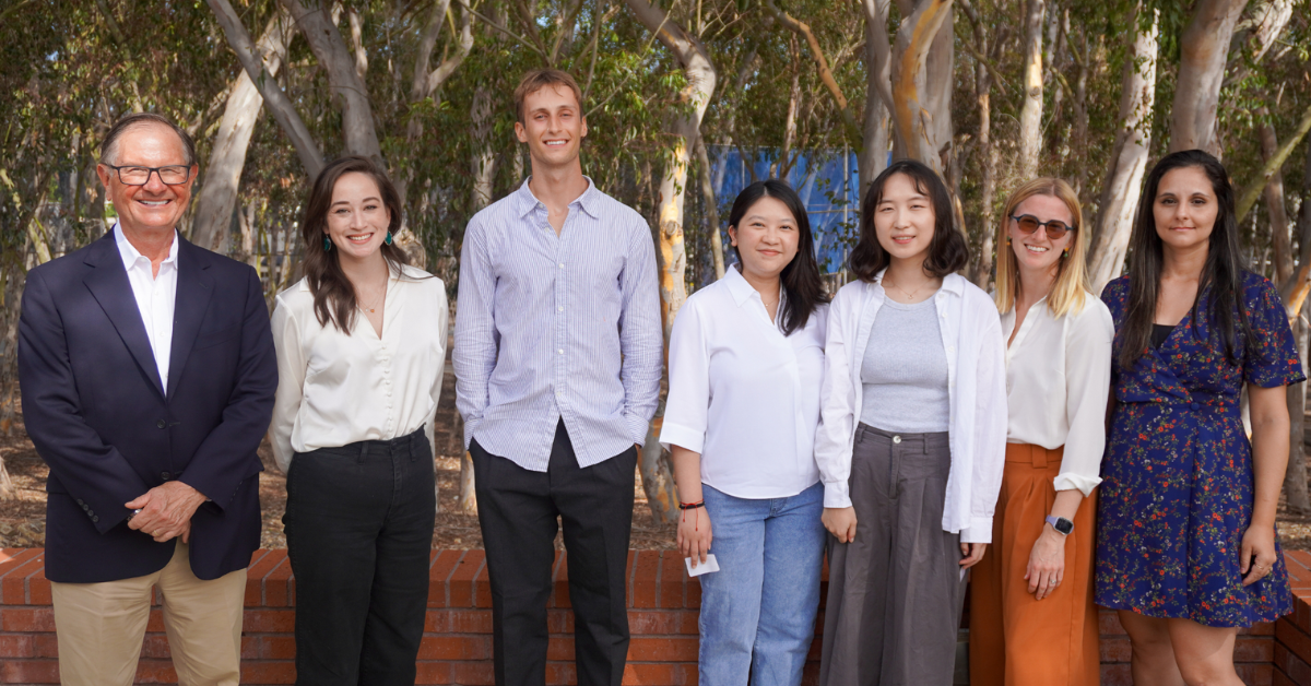 Seven individuals are standing in a line, should-to-should, outdoors, smiling and dressed in business casual attire, with trees in the background at the 2025 welcome event for the inaugural cohort of the Doctor of Philosophy in Public Health program.   