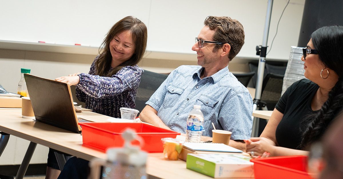 Three people smile and talk while seated in a classroom. 