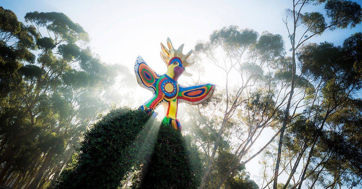 The sun shines behind a colorful bird statue surrounded by trees. 