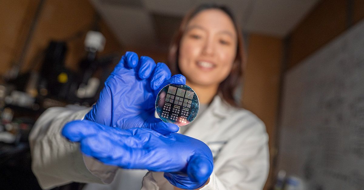 A small round metallic disk with tiny grid patterns is held by the gloved hand of a woman wearing a lab coat.