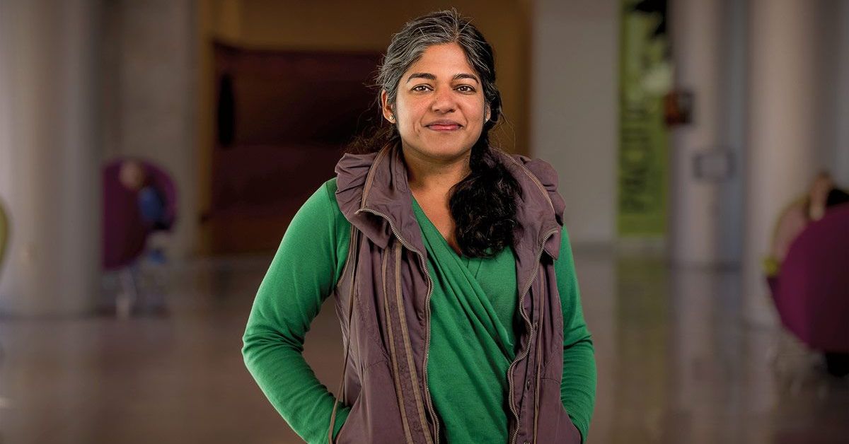 A standing woman in a green top and brown vest faces the camera with her hands in her pockets