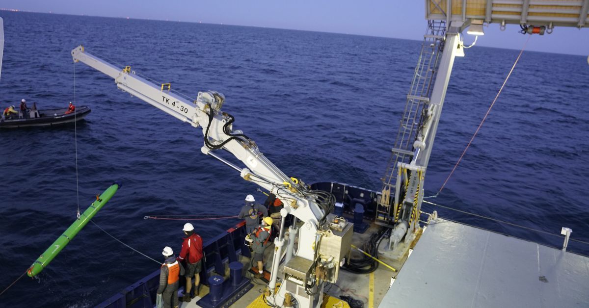 View of ship out to sea, a crane lowering a long slim green vessel into the water. 