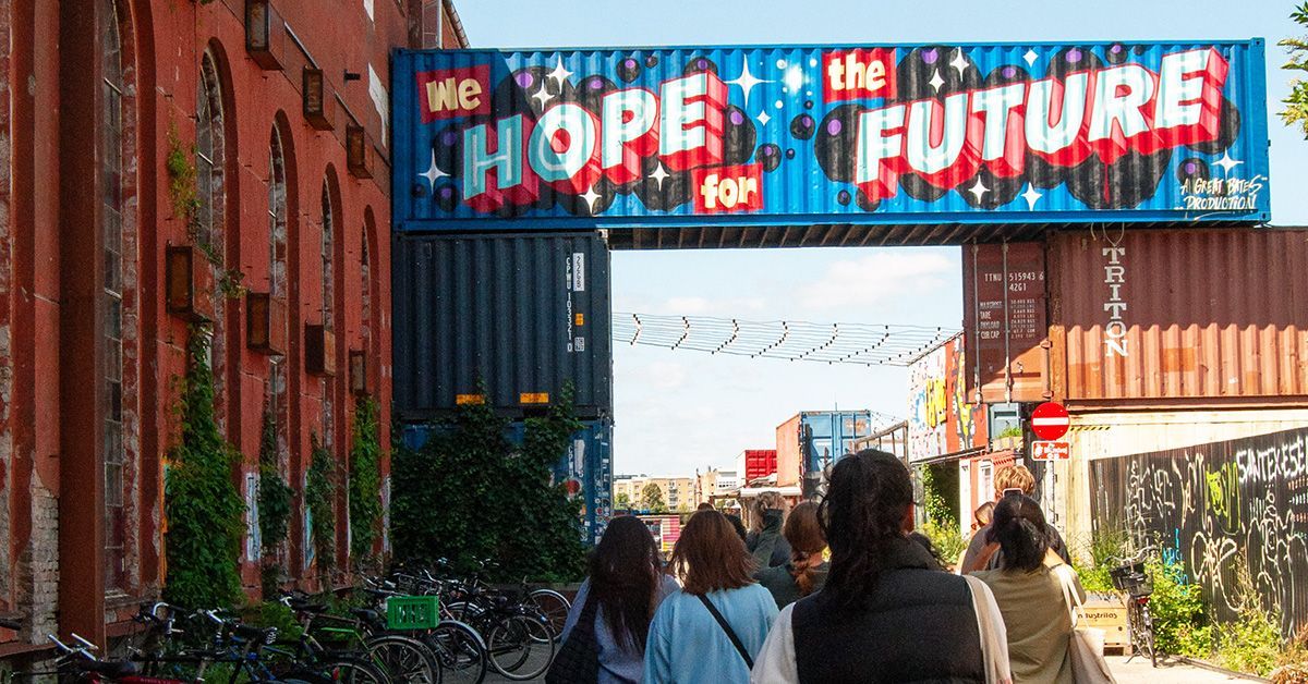A group of people walk into an outdoor market built of shipping containers. One of the containers is painted with a mural that says 
