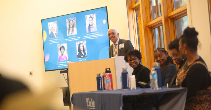 Four people sit at a long table facing an audience while a moderator looks on