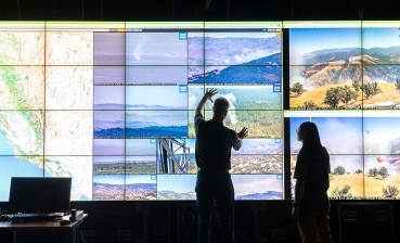 Two figures shown in shadow look and gesture toward a wall sized projection of camera images. 