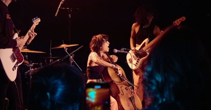 A seated cellist plays in a spotlight with a guitarist standing on her left and a bassist standing on her right.