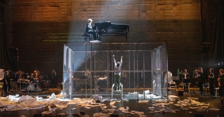 A piano is suspended above a stage. A person standing on a chair beneath the piano is is caged with chain-link fencing. Pieces of paper are scattered across the floor.